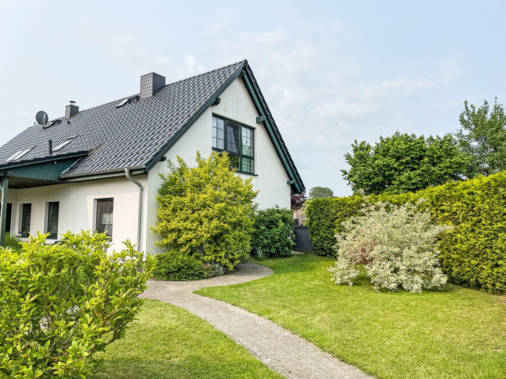 A white house with a sloped roof, a paved driveway, and a well-maintained garden with various shrubs and a pathway.