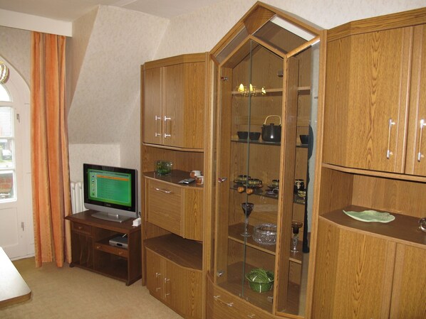 A room with a wooden cabinet containing kitchenware, a television on a stand, and a window with curtains.