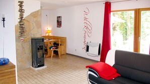 A living room with a black sofa, a red cushion, a stone wall, a fireplace, a wooden desk, and a window with a red curtain. - Waldhäuser by Interhome (Himmelpfort)