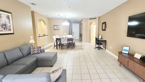 A living room with a grey sofa, a dining area with a table and chairs, a TV mounted on the wall, and a wooden sideboard with framed items. - Spacious 5-bedroom house in delightful Kissimmee with AC, WiFi (Kissimmee)