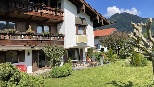 Ein zweistöckiges Gebäude mit Balkon, Garten und Bergen im Hintergrund.