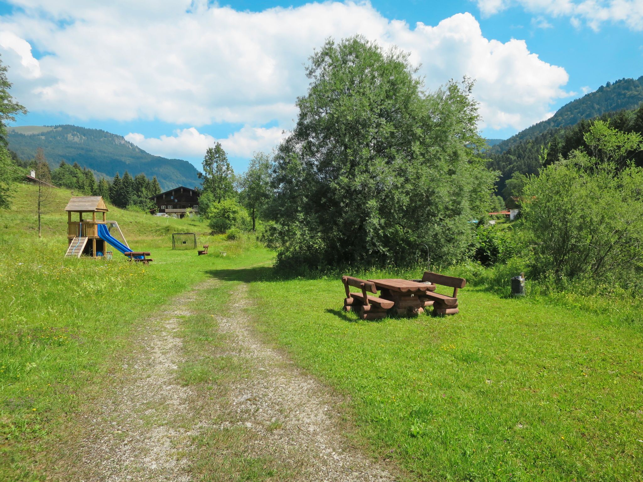 Un champ verdoyant avec une table de pique-nique, une aire de jeux avec un toboggan et, au fond, une cabane en bois.