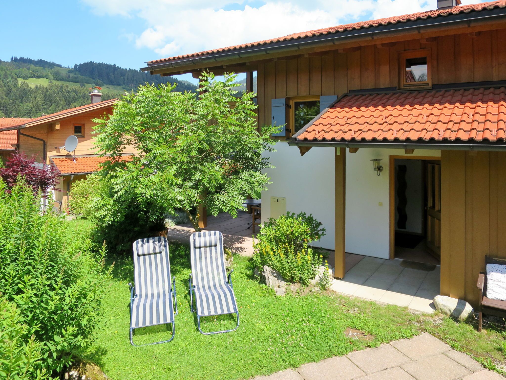 A two-story chalet with a red-tiled roof, a wooden exterior, and a garden with two striped lounge chairs.