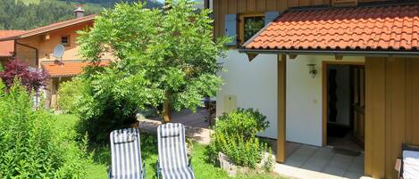 A two-story chalet with a red-tiled roof, a wooden exterior, and a garden with two striped lounge chairs.