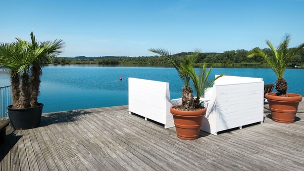 Een houten terras met witte lounge fauteuils en potpalmen, met uitzicht op een rustig meer.