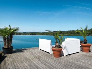 Eine Holzterrasse mit weißen Liegestühlen und Topfpalmen mit Blick auf einen stillen See.