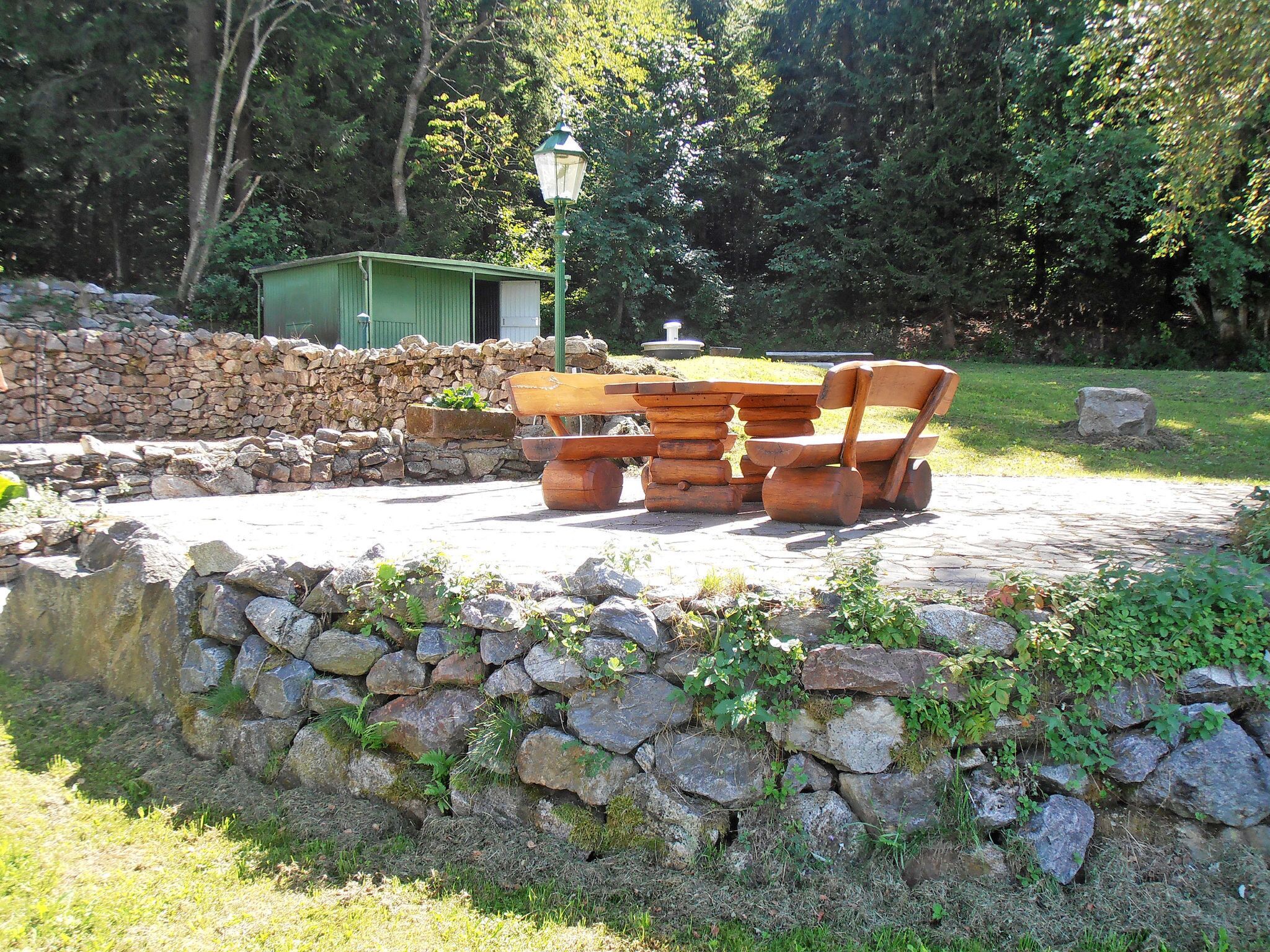 Un espace extérieur aménagé dans un style rustique, avec un mur en pierre, des bancs en bois et un abri de jardin vert, le tout situé dans un parc arboré.
