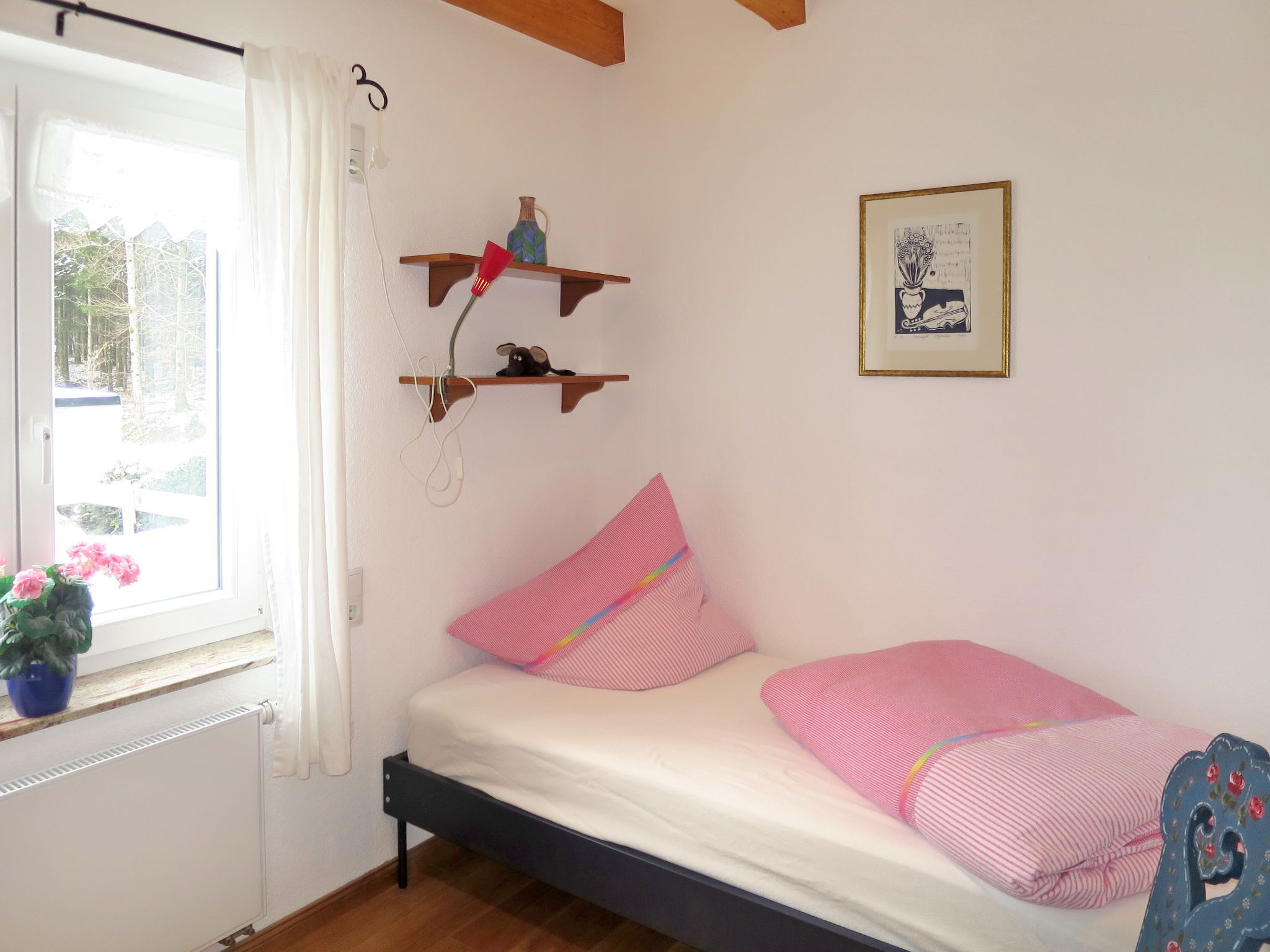A small bedroom with a single bed, a window with curtains, a radiator, and two wooden shelves with items on them.