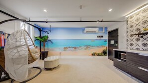 A room with a beach mural, a swing chair, a potted plant, a cushioned bench, and a modern kitchen area. - Westlake Hotel - The Beach (Etobicoke)