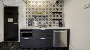 A compact kitchen with a dishwasher, a coffee machine, and a microwave. There is a black door with a handle, a small shelf with decorative items, and a patterned backsplash. - Westlake Hotel - The Beach (Etobicoke)