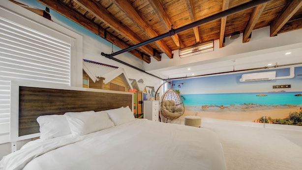 A bedroom with a large bed, wooden ceiling beams, a beach mural, and a hanging chair. - Westlake Hotel - The Beach (Etobicoke)