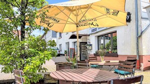 Een terras buiten met houten tafels en stoelen, een gele parasol en een gebouw met een bord waarop 