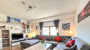 Een woonkamer met een grijze bank, een houten salontafel, een tv op een houten dressoir en een raam met gordijnen.