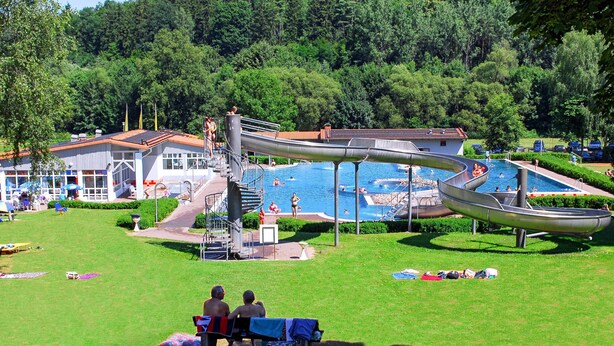A swimming pool with a water slide, surrounded by a grassy area and trees.