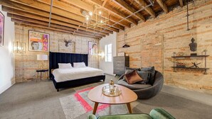 A room with exposed brick walls, a bed with a dark headboard, a grey sofa, a round coffee table, and a green armchair.