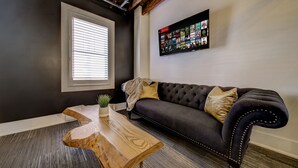 A modern living room with a dark gray wall, a tufted sofa with decorative pillows, a wooden coffee table with a plant, and a mounted TV displaying various channels. - Westlake Hotel - The Noire Suite (Etobicoke)
