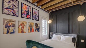 A bedroom with a bed, a green chair, and a wall with several framed artworks. - Westlake Hotel - The Art Suite (Etobicoke)