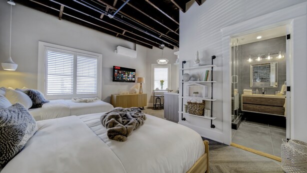 Une chambre moderne avec deux lits, un bureau et une salle de bain visible à travers une porte ouverte.