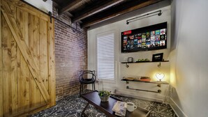 Une pièce chaleureuse avec une porte en bois, un mur en briques, un sol à motifs noir et blanc, une petite table avec une plante, une tasse de café et un écran Netflix fixé au mur.