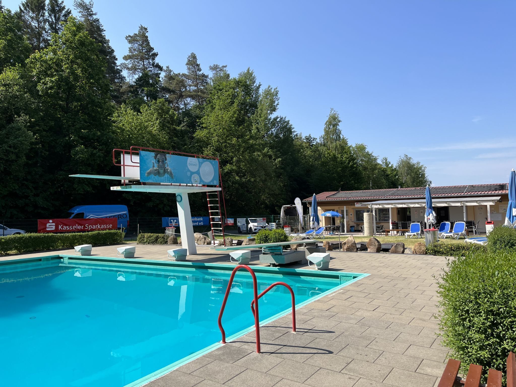 Une piscine dotée d’un plongeoir, de chaises longues et d’un bâtiment avec des espaces assises extérieurs.