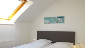 Un dormitorio con tragaluz, cama con ropa de cama blanca, mesita de noche con lámpara y un cuadro en la pared.