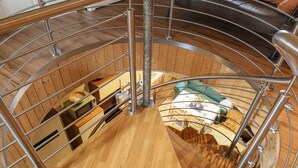 Una escalera en espiral con escalones de madera y barandillas de metal, que conduce a un nivel inferior con zona de comedor y sofá.