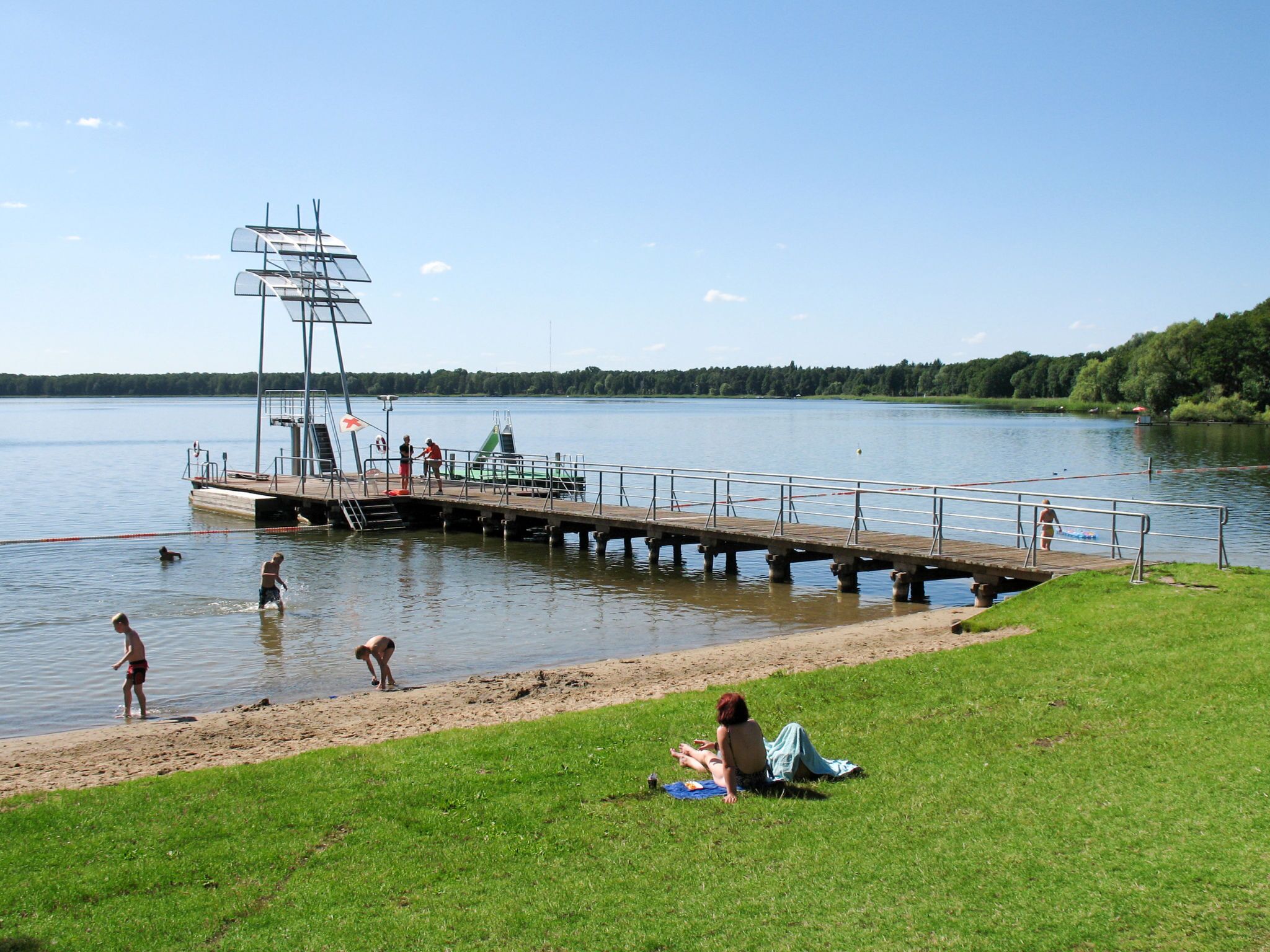 Une jetée au bord du lac, avec un poste de surveillance pour les sauveteurs, un petit bateau et des personnes pratiquant des activités nautiques.