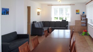 Un salón con sofá, una mesa de comedor y sillas. Hay una ventana con cortinas, una estantería y un cuadro en la pared.