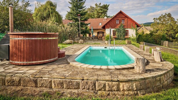 Zwembad omgeven door stenen trappen, een houten jacuzzi, en een huis met een rode dak en een stenen schoorsteen.