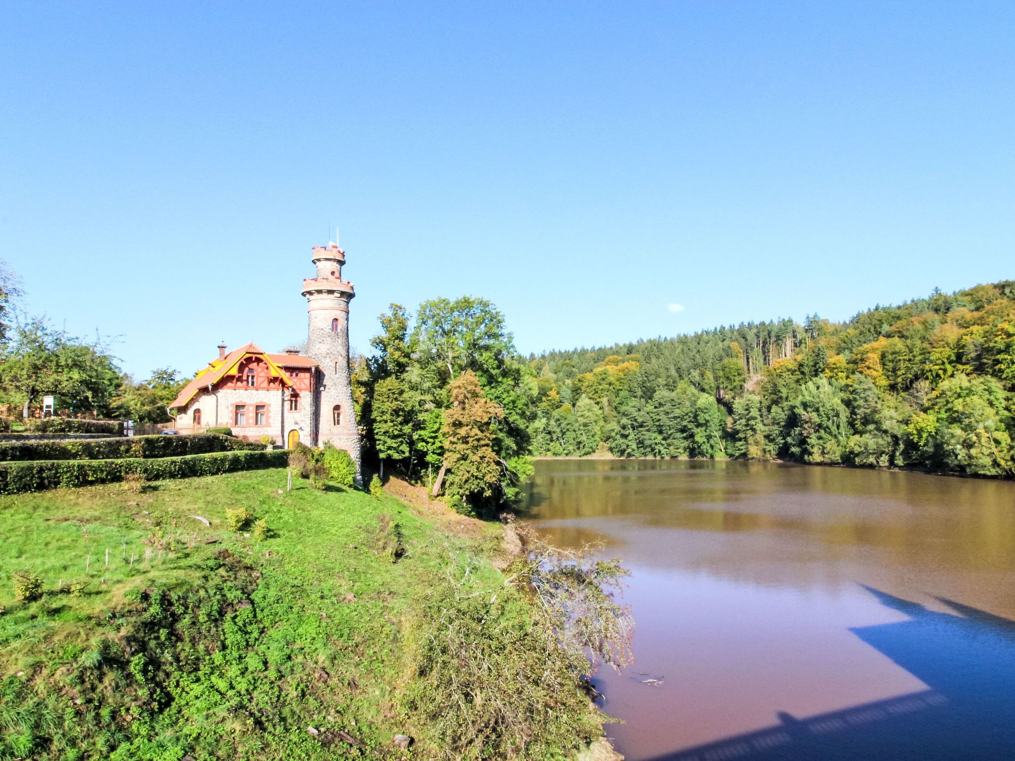 A historic stone building with a tower, situated by a calm river, surrounded by lush greenery and trees.