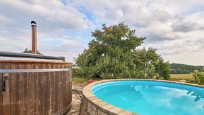 A wooden hot tub adjacent to a swimming pool with a stone border, surrounded by trees and a cloudy sky.