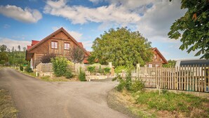 A rural road leading to a wooden house with a red roof, surrounded by a wooden fence and greenery.