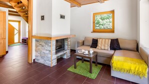 Un salon chaleureux avec une cheminée en pierre, des poutres apparentes en bois au plafond, un canapé et une fenêtre donnant sur la forêt.