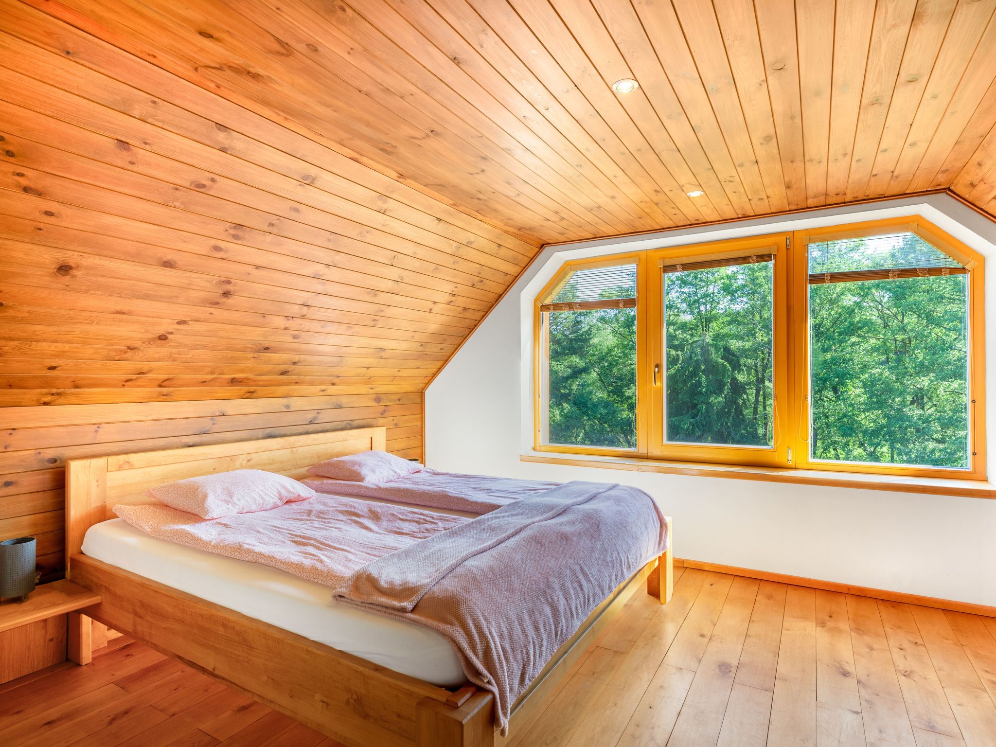 A bedroom with a wooden ceiling, a large window with a view of trees, a bed with pillows and a blanket, and a bedside table.