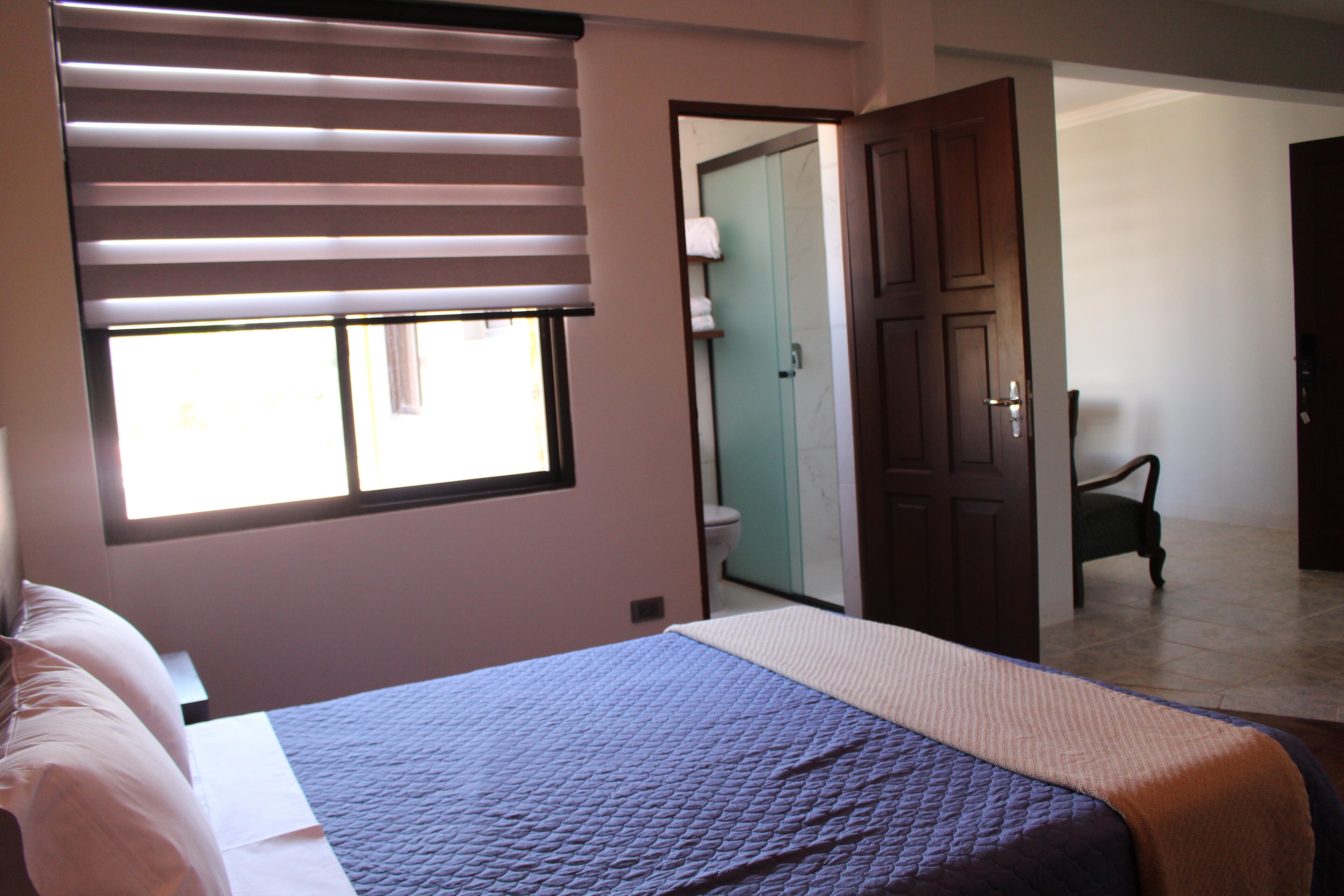 A bedroom with a bed, a window with blinds, a door leading to a bathroom, and a chair in the corner.