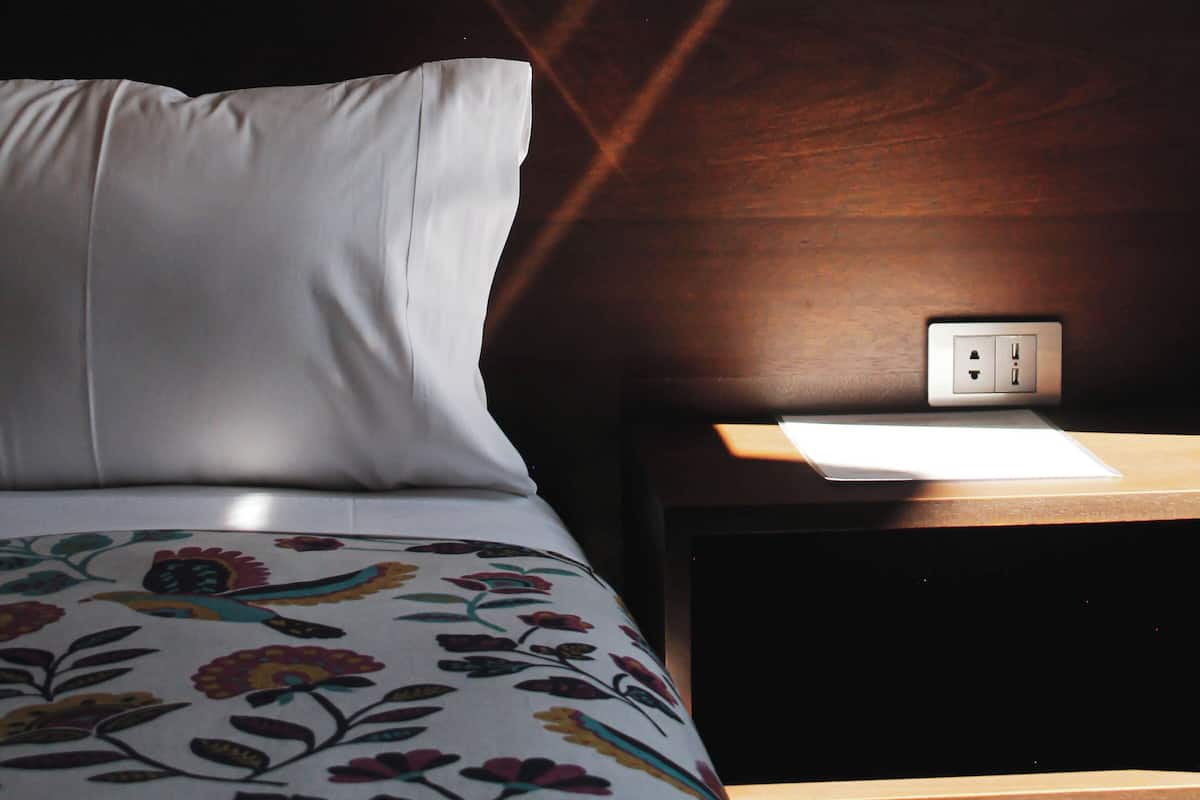A bed with a floral bedspread, a bedside table with a lamp, and a wall socket.