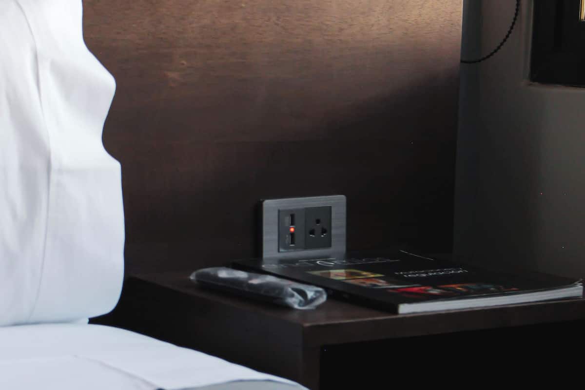A hotel room with a neatly made bed, a bedside table with a power outlet, a book, and a remote control.