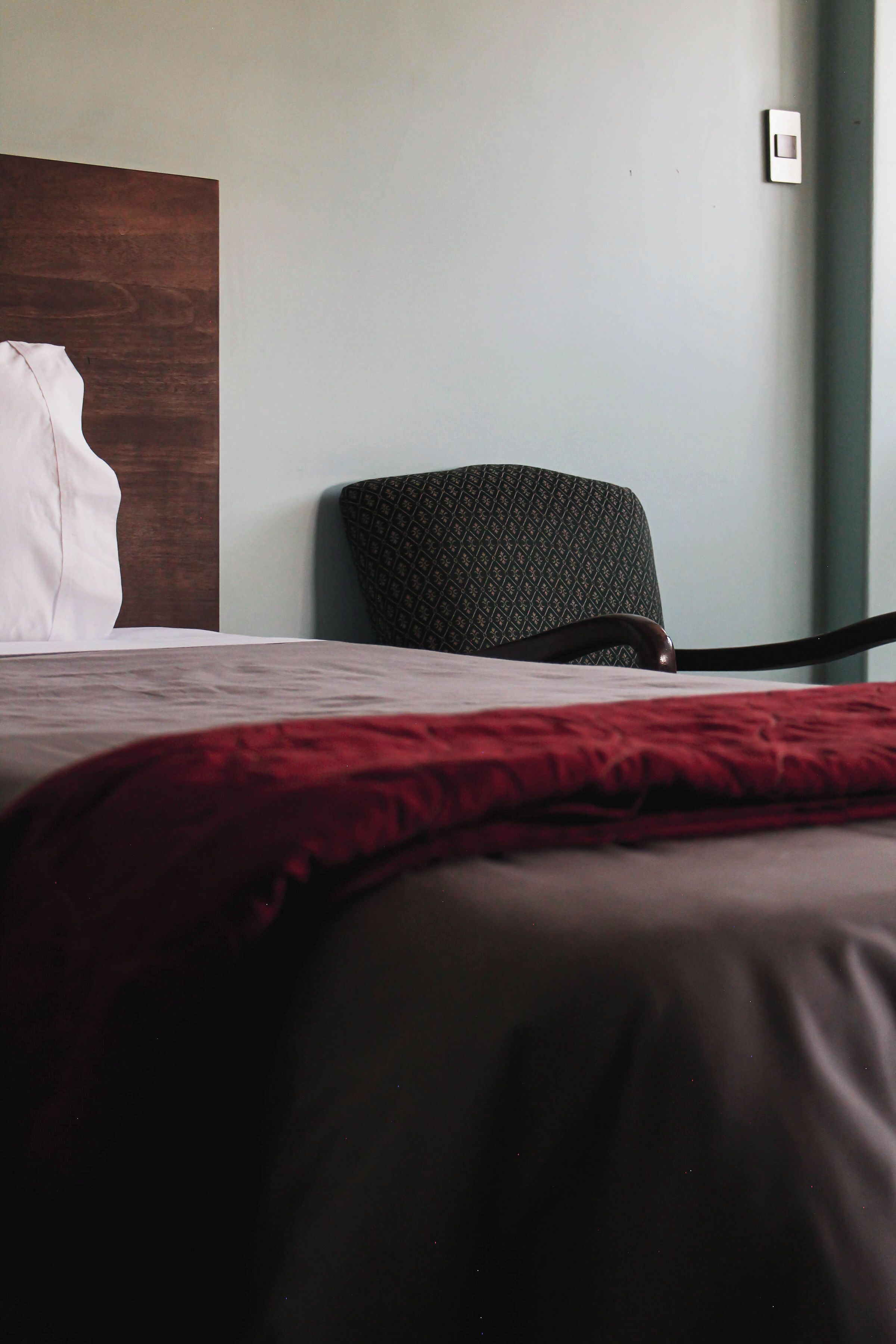 A bed with a red blanket, a wooden headboard, and a patterned chair against a plain wall.