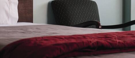 A bed with a red blanket, a wooden headboard, and a patterned chair against a plain wall.