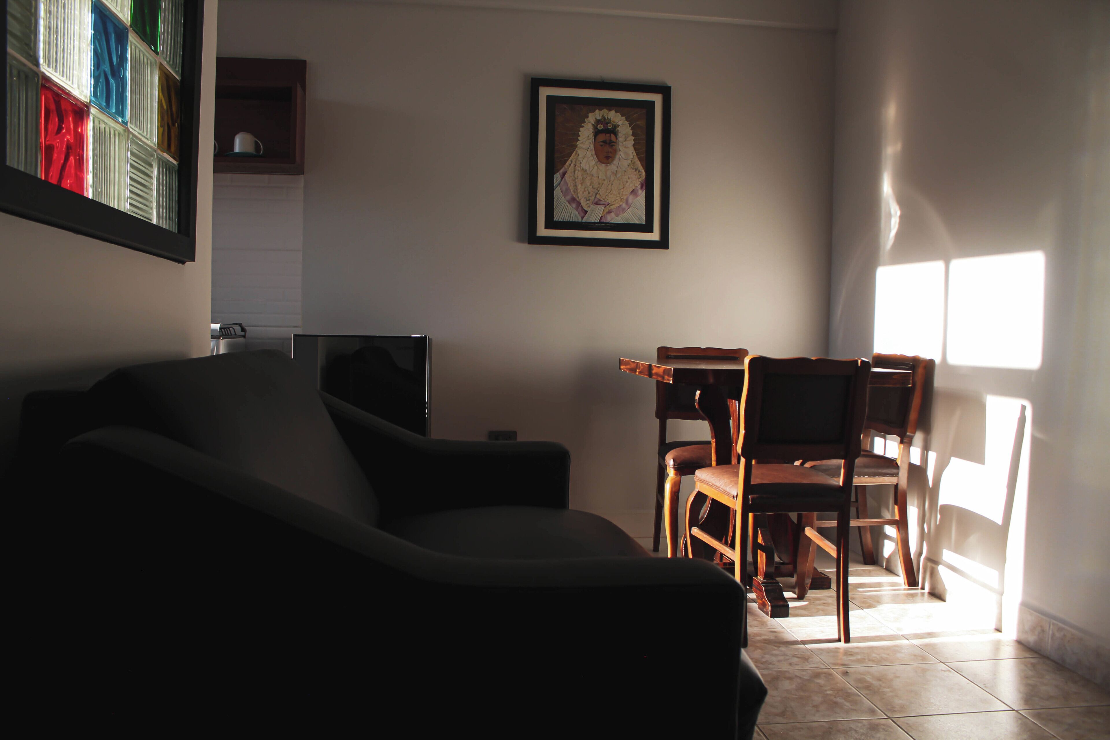 A living room with a sofa, a TV, and a dining table with chairs. There is a framed picture on the wall and a window providing natural light.
