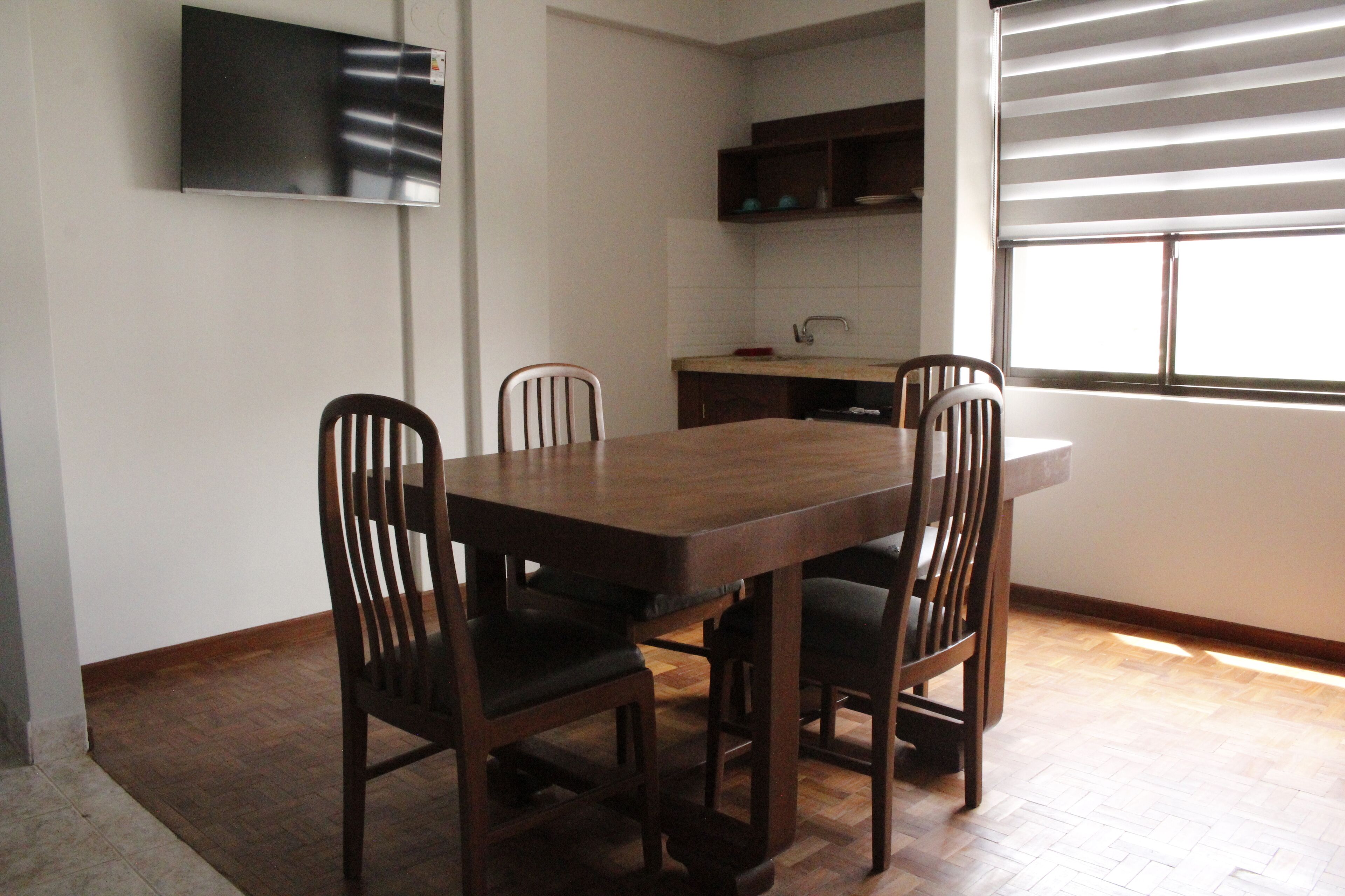 Un comedor con una mesa de madera y cuatro sillas, una cocina con fregadero y armarios, y una ventana con persianas.