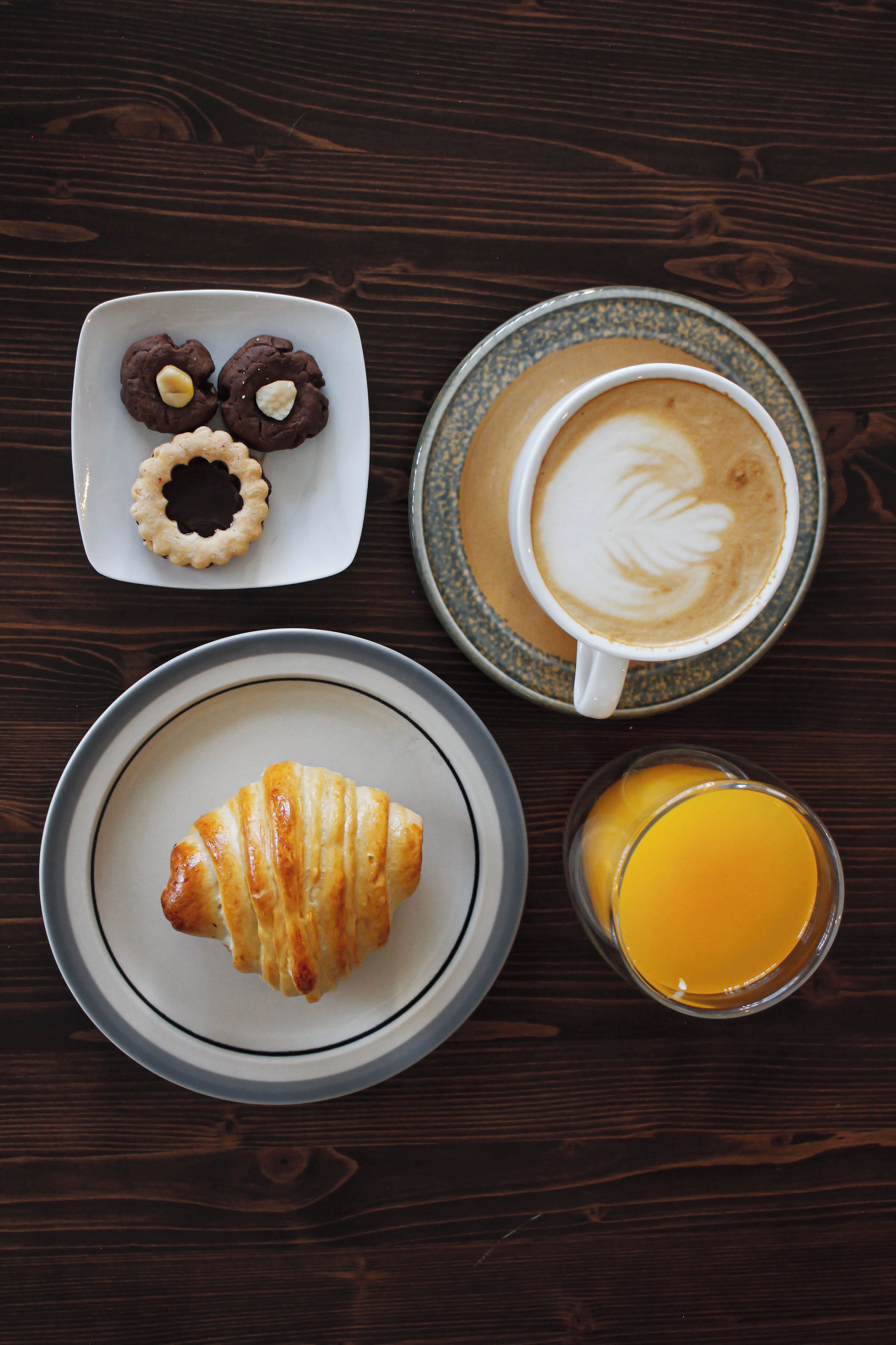 Una mesa de madera con un plato de cruasanes, un vaso de zumo de naranja y una taza de café con arte latte.