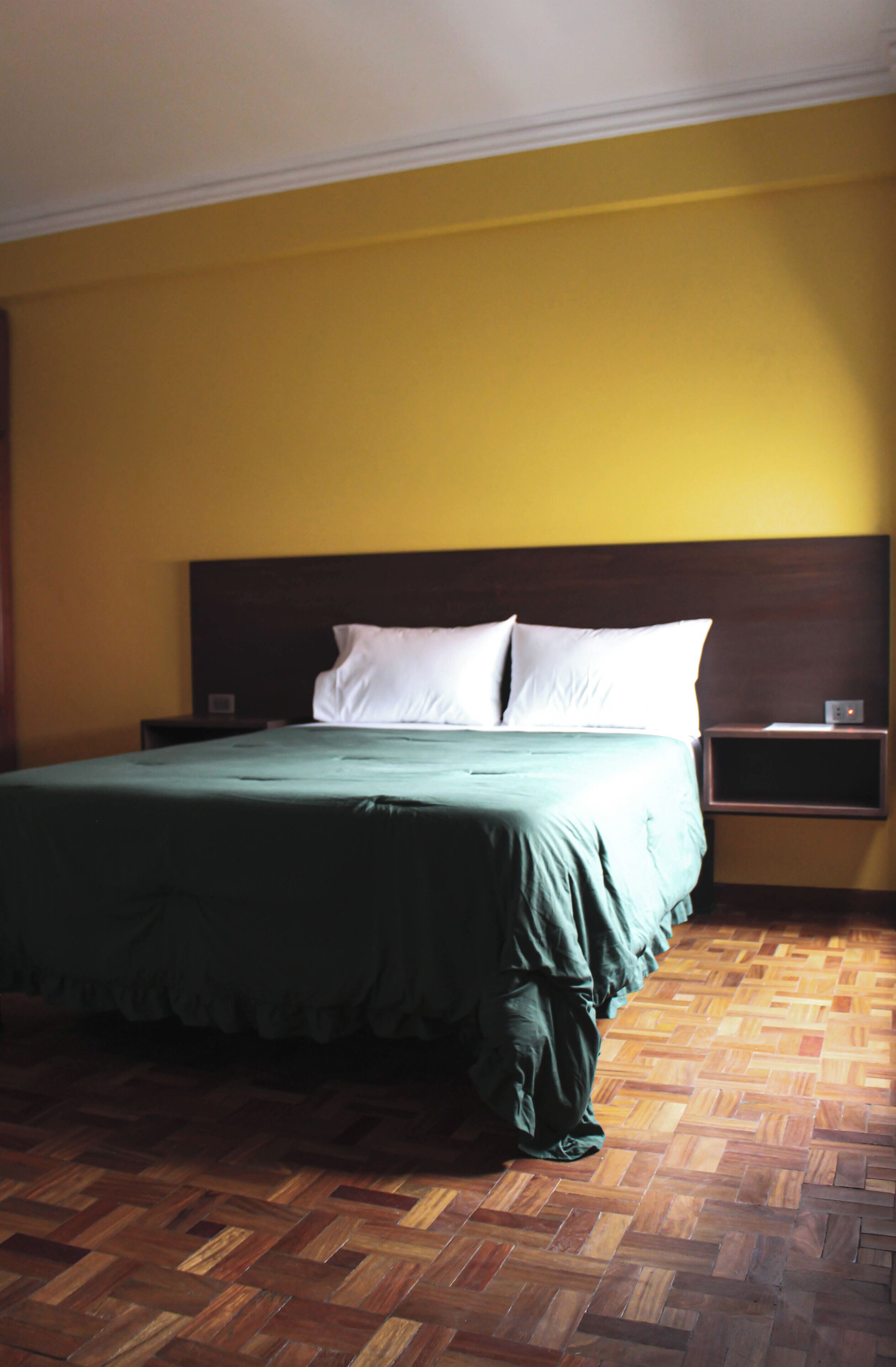 Una habitación de hotel con suelo de parqué de madera, colcha verde y pared amarilla.