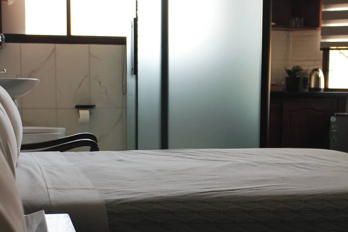 A hotel room with a bed, a bedside table, a window, and a kitchenette area.