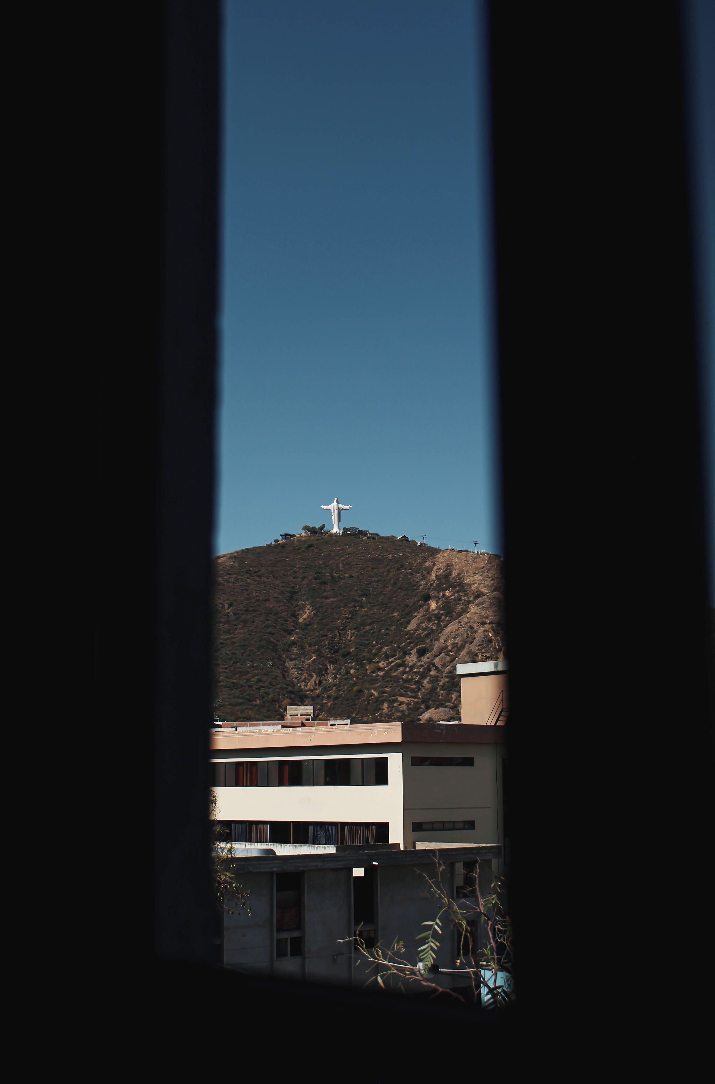 A view of a mountain with a statue on top, buildings in the foreground, and a clear sky.