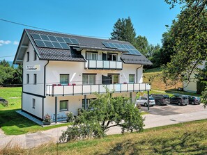 Un edificio a due piani con pannelli solari sul tetto, un balcone e un'area di parcheggio con auto.
