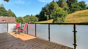 Una terraza de madera con dos sillas rojas y una barandilla de vidrio que da a un lago, rodeada de árboles y un paisaje montañoso.