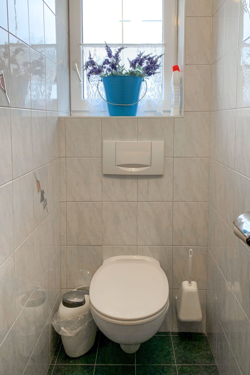 Un baño pequeño con inodoro, un cubo de basura, una ventana y una maceta azul con flores, además de un dispensador de papel higiénico.