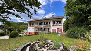 A two-story building with a garden in front, including a circular stone planter with plants and a small pond. - Apartment Meran by Interhome (Pörtschach am Wörthersee)