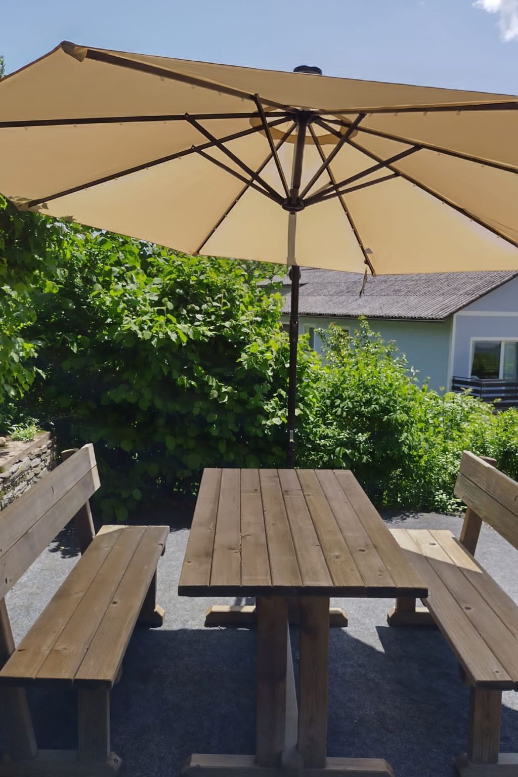 Et træborde med plads til picnic, overdækket af en stor beige parasol, omgivet af grøn vegetation og et hus i baggrunden.
